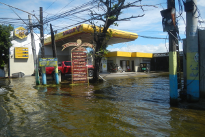 Flutstraße mit parkenden Autos in der Nähe einer Tankstelle, Wasser erreicht Strommasten und -leitungen, mit Texttafeln, Gebäuden, Bäumen und bewölktem Himmel im Hintergrund.