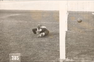 Ein Schwarz-Weiß-Foto eines Fussballspielers, der einen Ball vor einem Torpfosten mit Netz auf der rechten Seite fängt und unten Text.