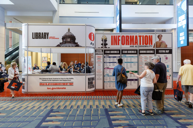 Eine Gruppe von Menschen auf dem Nationalen Buchfest, einige sitzen und einige stehen, vor einer Theke mit Bannern, einem Anzeigebildschirm, einer Treppe, einem Geländer und einer Decke mit Deckenleuchten.