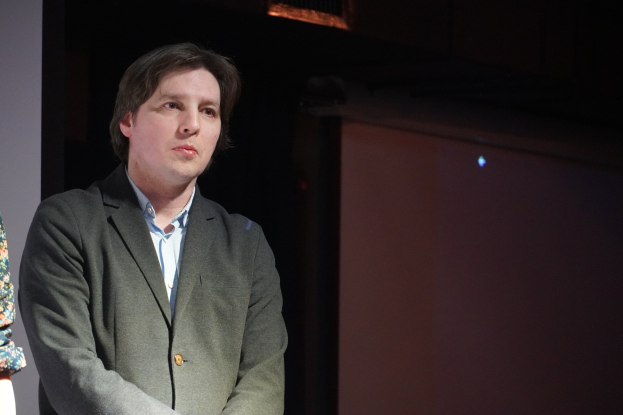 Zwei Menschen in formeller Kleidung stehen nebeneinander auf einer Bühne mit ernsten Gesichtern.