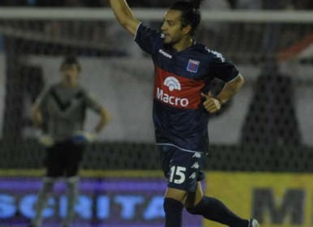 Ein Fußballer feiert auf dem Boden nach einem Tor, mit einer jubelnden Menge und einer Person im Hintergrund und einer Texttafel dahinter, alles mit einem unscharfen, energetischen Hintergrund.