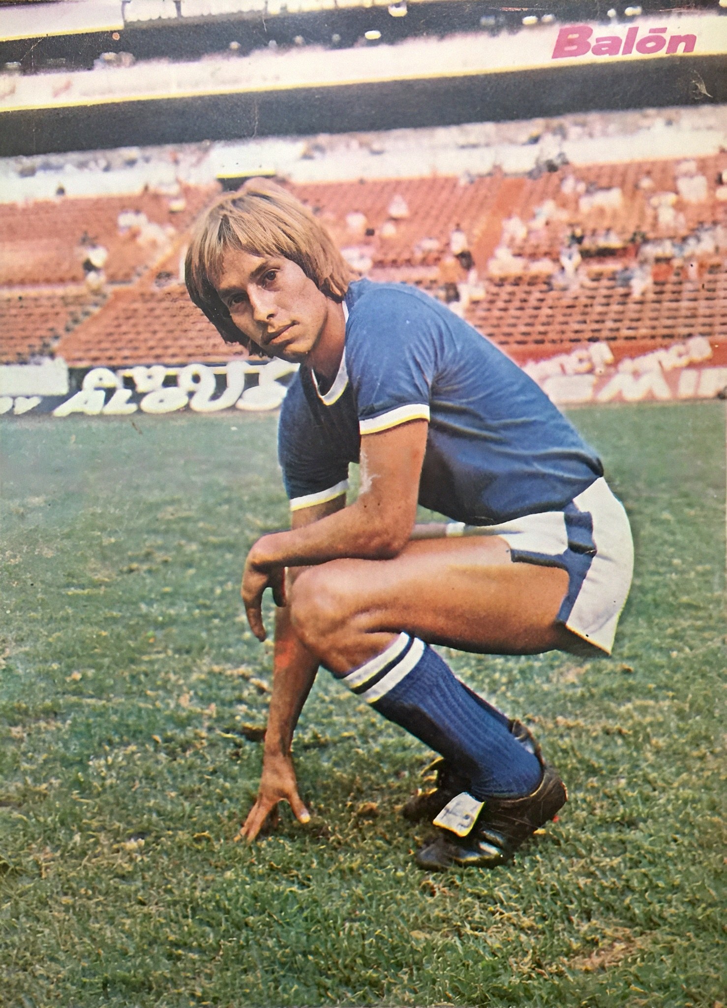 Ein Mann kniend auf einem Fußballfeld umgeben von Menschen auf Stühlen und Schildern mit Text, trägt Schuhe.