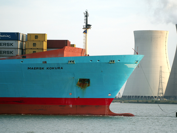Großes blaues und rotes Containerschiff mit gestapelten Cargo-Containern, das in der Nähe eines Atomkraftwerks segelt, mit einem Kühlturm im Hintergrund bei klarem Himmel.