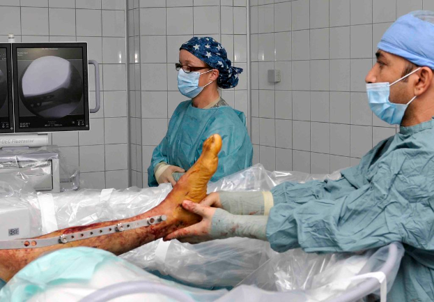 Eine Person in einer Maske und Kopfbedeckung hält ein Bein mit einem Stab und etwas Blut daran, das auf einem Bett mit weißen Fliesenwänden und Monitoren liegt, die ein Röntgenbild anzeigen, während eine andere Person in einer Maske und Kopfbedeckung auf der anderen Seite des Bettes steht.