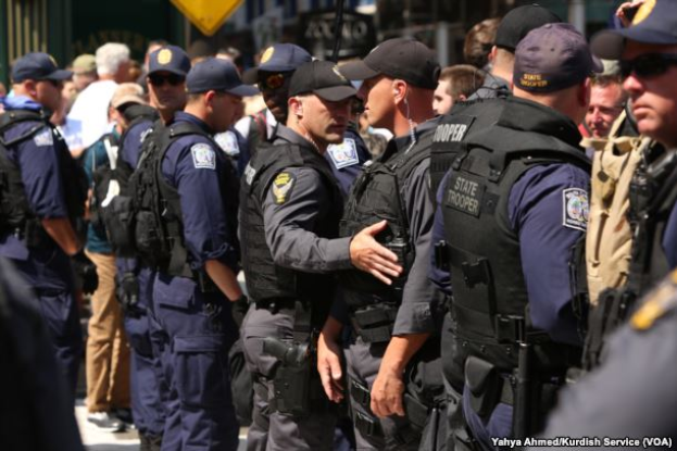 Eine Gruppe von Polizisten in Mützen, Taschen und Schutzbrillen, einige mit Waffen in der Hand, steht zusammen auf einer Straße mit Gebäuden, Schildern und Polen im Hintergrund und Text am unteren Rand des Bildes.