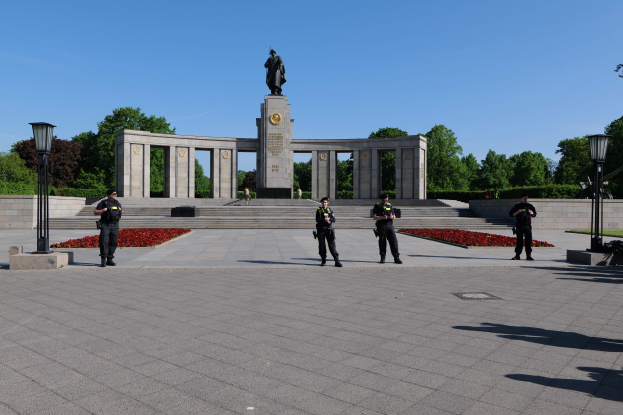 Eine Gruppe von Polizisten in schwarzen Uniformen, die Gewehre halten, steht vor einem Denkmal mit einer Statue darauf, umgeben von Laternenmasten, mit Stufen, die hinaufführen, und einem Hintergrund aus Bäumen unter einem klaren blauen Himmel.