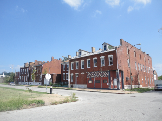 Ein Geb├Ąude mit Masten und Stra├čenlaternen, umgeben von B├Ąumen und Fahrzeugen auf beiden Seiten, mit einem Weg, Gras und einem Feuerl├Âscher unten und dem Himmel oben.