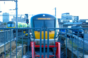 Zwei Züge auf parallelen Gleisen mit einem Zaun im Vordergrund, ein Auto auf der Straße rechts daneben, eine Signalleuchte auf einem Pfahl, Gebäude im Hintergrund und Himmel oben.