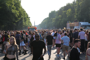 Eine große Menge Menschen, die eine von Bäumen gesäumte Straße entlanggehen, mit einem Turm im Hintergrund, Fahrzeugen mit Menschen auf der rechten Seite, wahrscheinlich beim Christopher Street Day in Berlin unter einem klaren blauen Himmel.