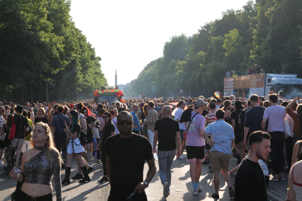 Eine große Menge Menschen, die eine von Bäumen gesäumte Straße entlanggehen, mit einem Turm im Hintergrund, Fahrzeugen mit Menschen auf der rechten Seite, wahrscheinlich beim Christopher Street Day in Berlin unter einem klaren blauen Himmel.