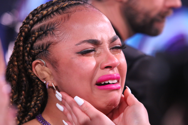 Eine Frau mit Zöpfen und pinkem Lippenstift weint im Vordergrund, während im Hintergrund einige Menschen und ein unscharfer Hintergrund zu sehen sind.