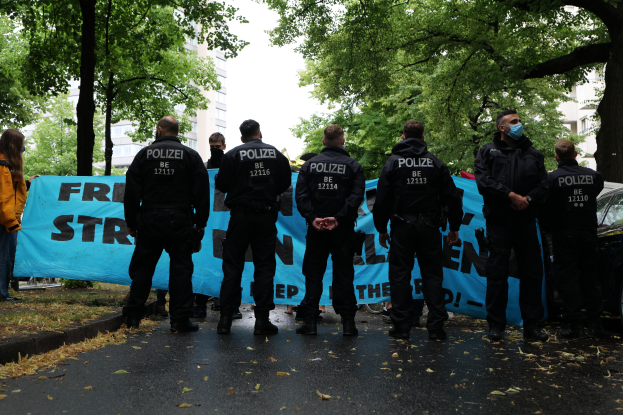 Eine Gruppe von Polizisten steht vor einer blauen Fahne mit Text, umgeben von Bäumen und Gebäuden, mit einem geparkten Auto auf der rechten Seite und einer Frau in einer Jacke auf der linken Seite, in Berlin, Deutschland.