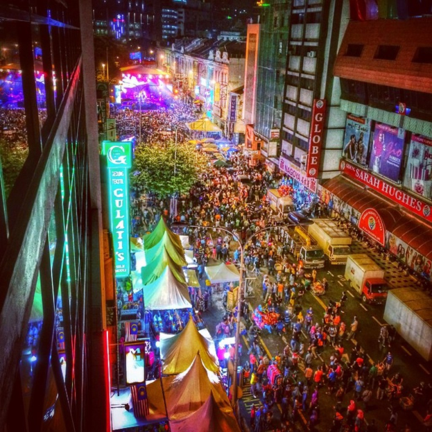 Eine volle Karnevals-Szene mit Zelten, Geschäften, Gebäuden und Bannern, mit hell erleuchteter Fläche im Hintergrund.