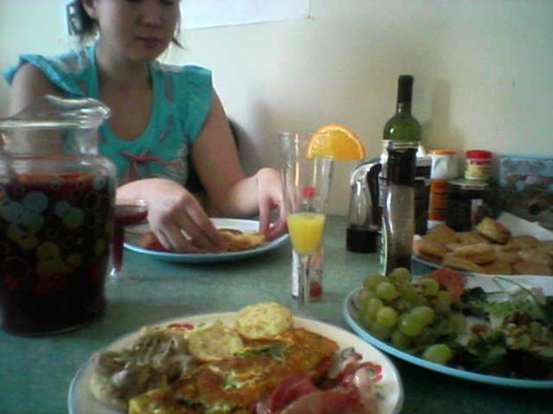 Eine Frau sitzt auf einem Stuhl mit einem Teller voller Essen, darunter Trauben, und verschiedenen Gegenständen wie einem Glas, einer Flasche und einer Schachtel in der Nähe.