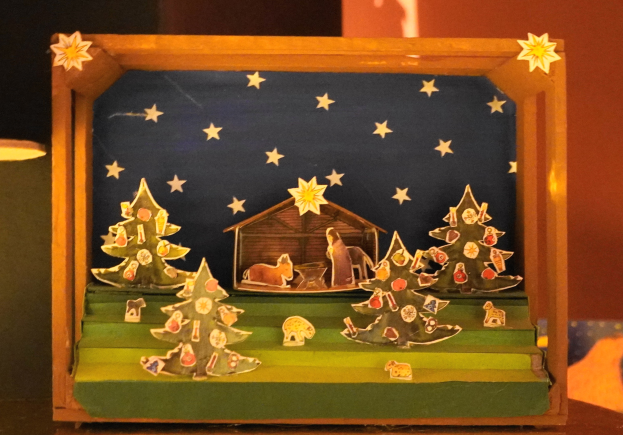 Eine hölzerne Krippe auf einem Tisch, mit einer Lampe links und einer Wand im Hintergrund, geschmückt mit Weihnachtsbäumen, Tieren und einem Haus.