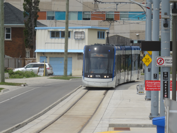 Eine Straßenbahn fährt durch eine Straße mit hohen Gebäuden, mit Masten, Schildern, einem Mülleimer, einem Auto und einem Baum.