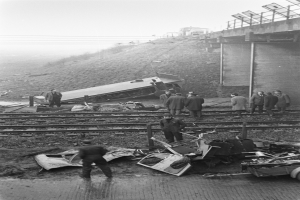 Ein Schwarz-Weiß-Bild eines Zugunglücks mit Menschen um das Wrack herum, ein Fahrzeug im Vordergrund und eine Brücke, Masten, Drähte und Himmel im Hintergrund.