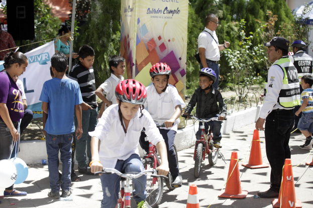 Gruppe von Kindern, die auf der Straße mit Verkehrskegeln Fahrrad fahren, einige tragen Helme, andere stehen in der Nähe, mit einem Banner, Bäumen und Gebäuden im Hintergrund.