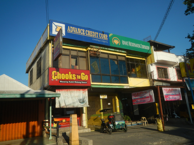 Ein Gebäude mit einer Tafel, auf der "Advance Credit Corp" steht, auf einer Straße mit Fahrzeugen, Werbetafeln, Strommasten, Kabeln, Bäumen und einem klaren blauen Himmel.