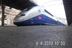 Ein Zug ist in der Mitte des Bildes mit einer Plattform und Text unten, Gebäuden und einem Dach links und Gebäuden, Dächern, Kabeln und Himmel oben.