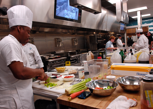 Eine Gruppe von Köchen in einer Küche, die auf einem Schneidbrett Lebensmittel vorbereiten, wobei ein Koch in der foreground in einem weißen Kleid und einer Mütze steht, ein Messer hält und von Utensilien, Schüsseln, Gemüse, einem Mobiltelefon, Flaschen und anderen Gegenständen umgeben ist, mit einem Fernseher und Deckenlampen im Hintergrund.