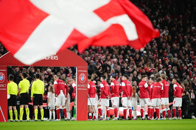Gruppe von Menschen auf einem Fußballfeld mit einer roten und weißen Flagge im Vordergrund, einem Bogen mit dem Text "Bayern München vs. Bayern München Wett-Tipps & Vorschau" im Hintergrund und einer großen Menge im Stadion.