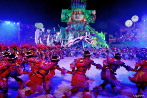 Eine Gruppe von Menschen in bunten Kostümen tanzt auf einer Bühne vor einem sitzenden Publikum, mit Lichtern, Pfählen, Ballons und einem Bildschirm, der eine Person in den Hintergrund zeigt, während des Karnevals in Rio de Janeiro 2014.
