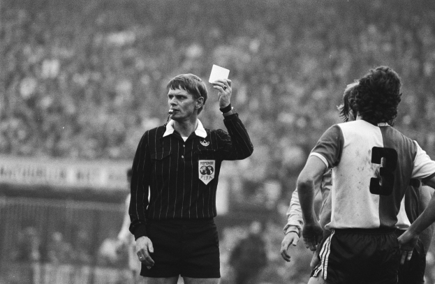 Schwarzes-weißes Bild eines Schiedsrichters mit roter Karte, mit einer Menge von Menschen in Fußballuniformen hinter einem Zaun.