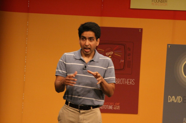 Ein Mann steht auf einer Bühne und spricht, mit einer Wand voller Plakate im Hintergrund.