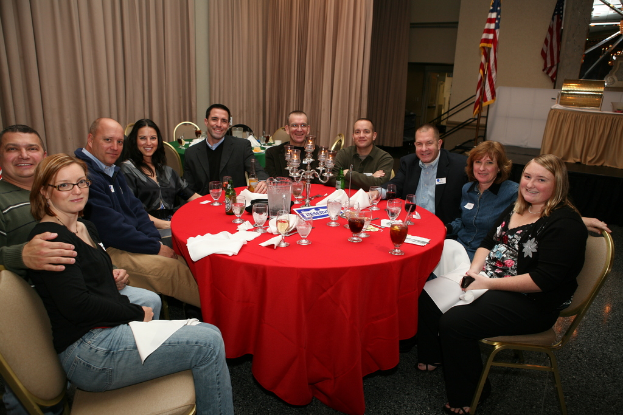 Eine Gruppe von Menschen mit einem Glas Wein, Papieren, Gläsern und Flaschen auf dem Tisch, einer Flagge auf der rechten Seite und Vorhängen an der Wand.
