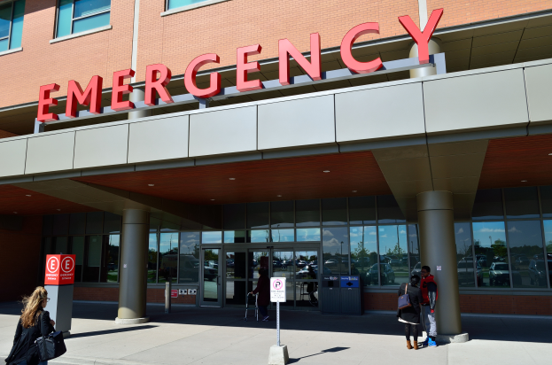 Eine Gruppe von Menschen vor einem gl"asernen Notfallgebäude mit S"ulen, einem Namensschild, Schildern auf Pfosten und einem Müllcontainer.