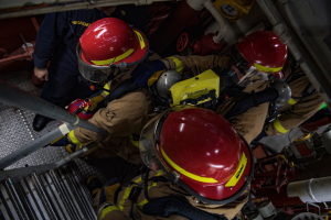 Eine Gruppe von Feuerwehrleuten in Helmen und Schutzausrüstung arbeitet an einem Feuerlöschfahrzeug, mit Rohren und anderen Gegenständen im Hintergrund, was darauf hinweist, dass sie aktiv ein Feuer bekämpfen.