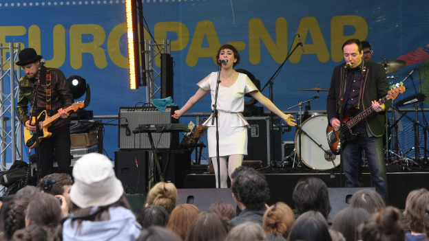 Ein Konzert mit einer Frau in einem weißen Kleid in der Mitte, zwei Männern, die Gitarren spielen, einer Menge unten und Lautsprechern im Hintergrund.