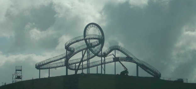 Eine Achterbahn auf dem Gipfel eines Hügels unter einem bewölkten Himmel, umgeben von Stützkonstruktionen.