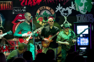 Drei M├Ąnner, die auf einer B├╝hne Gitarren spielen, mit einer Menge unten und einem Graffiti oder einer Wandmalerei im Hintergrund.