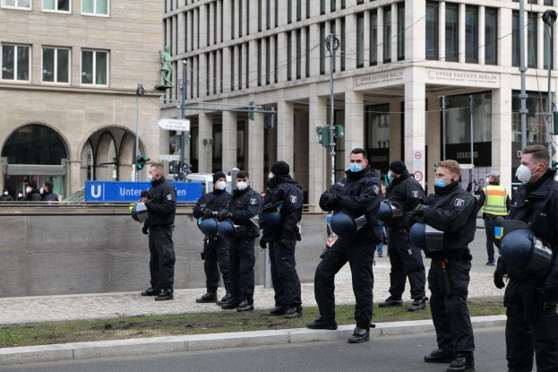 Eine Gruppe von Polizisten in schwarzen Uniformen und Masken steht vor einem Gebäude mit Glasfenstern und Säulen in Berlin, Deutschland, einige halten Helme, mit Laternen, Verkehrsampeln, Schildern mit Text, einer Statue und anderen Menschen im Hintergrund.