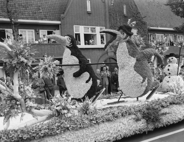 Schwarzes und weißes Foto eines Paradewagens, der mit Blumen und Pinguinfiguren geschmückt ist, mit Menschen auf der Straße und Gebäuden mit Fenstern im Hintergrund.