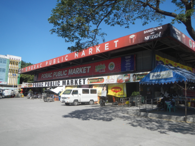 Ein öffentlicher Markt mit einem vor ihm geparkten Transporter, umgeben von Gebäuden, Geschäften, Schildern, Kraftfahrzeugen auf der Straße, Menschen unter einem Zelt sitzend, Geländern, Bäumen und einem klaren blauen Himmel.