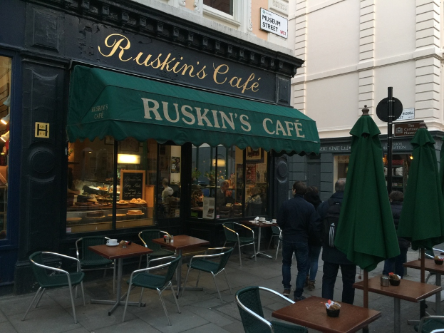 Eine Gruppe von Menschen vor einem Café mit einer Tafel, auf der 'Ruskin's Cafe' steht.