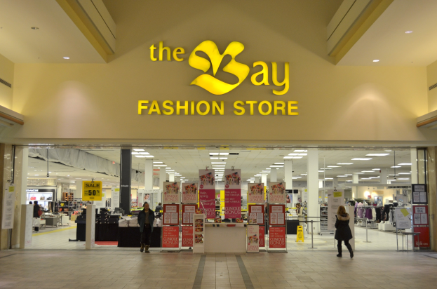 Außenansicht eines Bay Fashion Stores in einem Einkaufszentrum mit Passanten, Wandtafeln, Säulen, Deckenleuchten und Hintergrundauslagen.