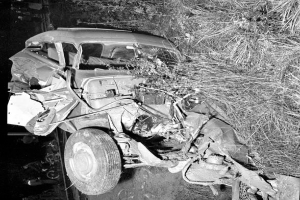 Ein Schwarz-Weiß-Foto eines schwer beschädigten Autos am Straßenrand mit einer eingedrückten Front, zersplitterten Fenstern, einem Baum im Hintergrund und verstreuten Gegenständen rund um die Szene.