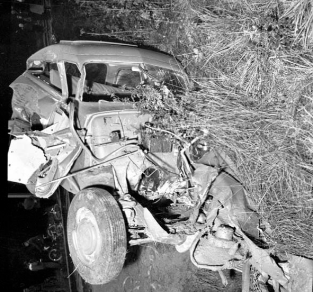 Ein Schwarz-Weiß-Foto eines schwer beschädigten Autos am Straßenrand mit einer eingedrückten Front, zersplitterten Fenstern, einem Baum im Hintergrund und verstreuten Gegenständen rund um die Szene.