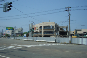 Eine Stadtstraße mit Fahrzeugen, Strommasten, Laternen und einer Ampel auf der linken Seite, Gebäude, Bäume und den Himmel im Hintergrund und ein Gebäude in derForeground, das abgerissen wird.