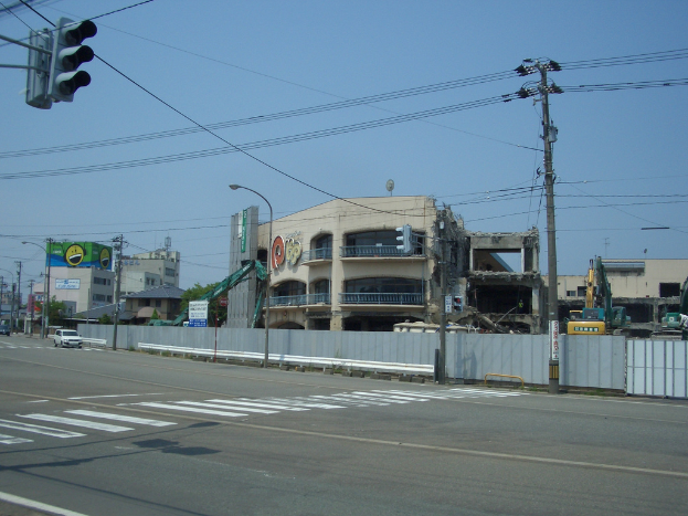 Eine Stadtstraße mit Fahrzeugen, Strommasten, Laternen und einer Ampel auf der linken Seite, Gebäude, Bäume und den Himmel im Hintergrund und ein Gebäude in derForeground, das abgerissen wird.