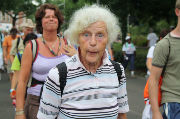 Eine öltere Frau mit einem Überraschten Gesichtsausdruck geht die Straße entlang, umgeben von Menschen, einige tragen Taschen, mit Bäumen, Gebäuden und einem klaren Himmel im Hintergrund.