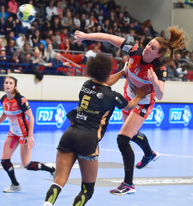 Eine Gruppe von Frauen spielt Handball auf einem Feld mit einem Ball in der Luft, während Zuschauer von Stühlen, Geländern und einer Wand im Hintergrund zusehen.