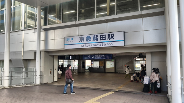 Eine Gruppe von Menschen, die durch einen Bahnhof geht, einige tragen Gepäck, mit einer Tafel mit Text, Pfeilern, Geländern, Glaswänden und Deckenlampen; ein Mann in der Mitte mit Schuhen.