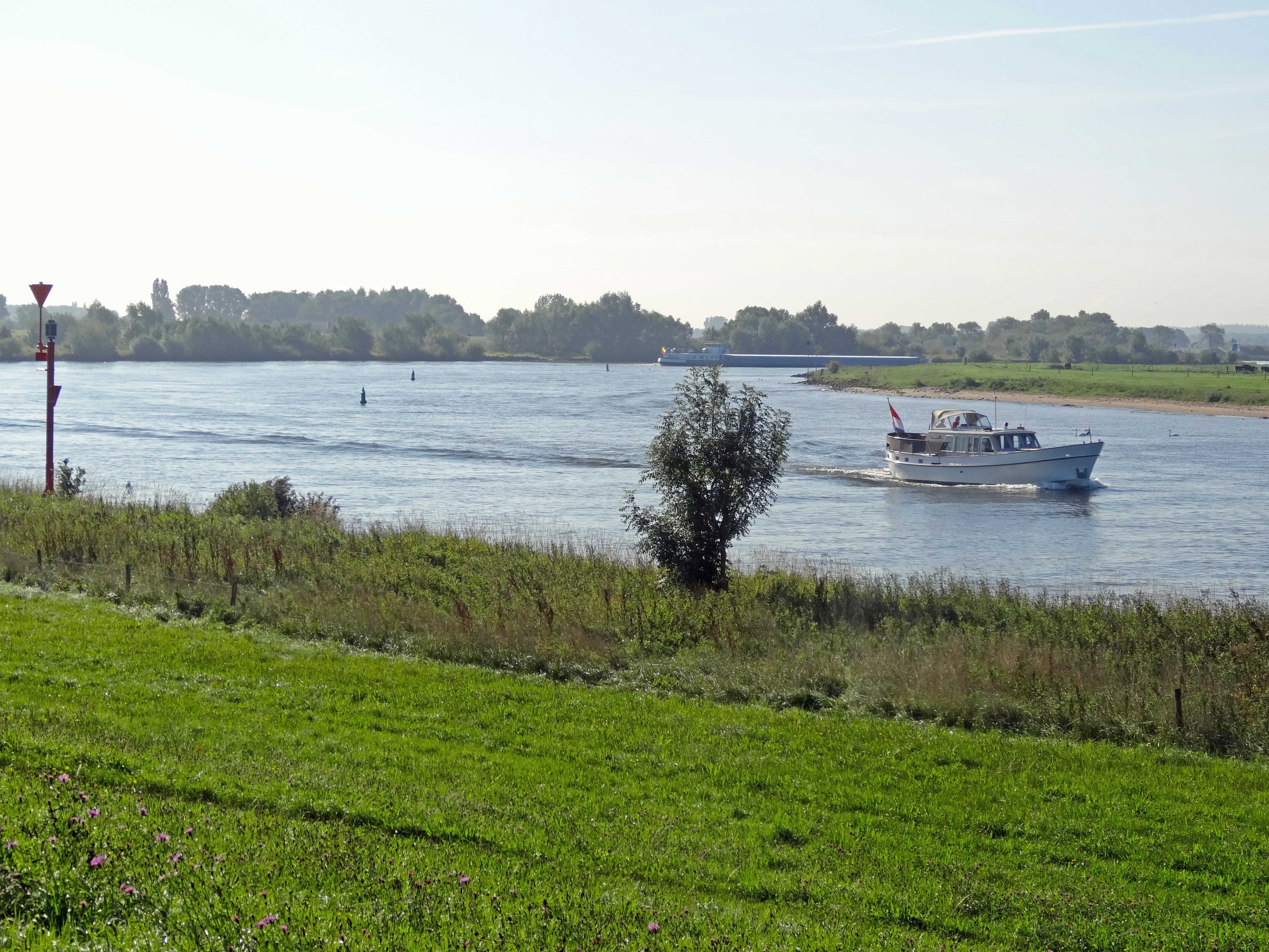 Ein Boot, das einen von Grünflächen, Pflanzen und Blumen gesäumten Fluss entlangfährt, mit Bäumen und einem blauen Himmel im Hintergrund.