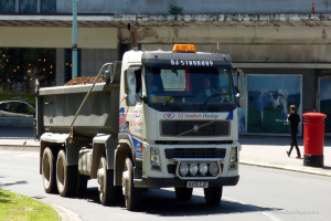 Ein Lastwagen fährt auf einer Straße mit Gebäuden im Hintergrund, einer Briefkasten, einer gehenden Person, einem Pfahl und Grasland.