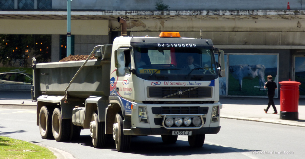 Ein Lastwagen fährt auf einer Straße mit Gebäuden im Hintergrund, einer Briefkasten, einer gehenden Person, einem Pfahl und Grasland.